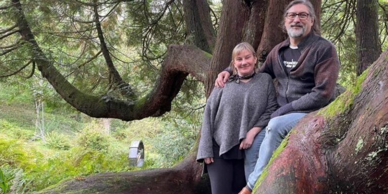 Photo of middle aged couple stood in the woods, in front of a large tree