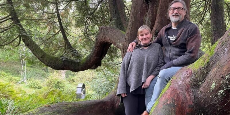 Photo of middle aged couple stood in the woods, in front of a large tree